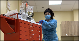 A healthcare workers with black curly hair wearing a mask in a clinic pushing a red medical cart