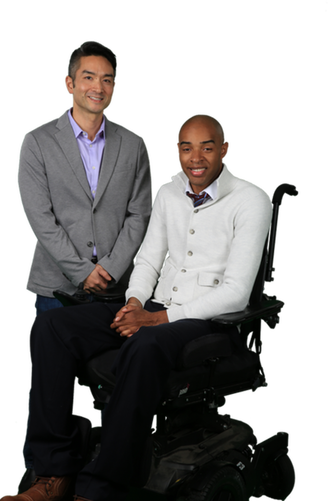 Two smiling professional next to each other one is standing wearing a gray blazer and the other is on a wheelchair with a with a button up